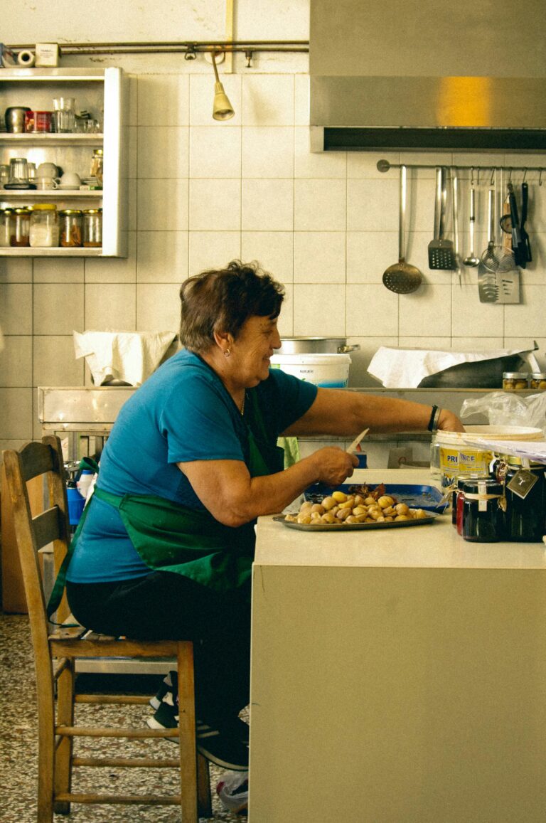 Senior woman seated in a Pilio kitchen, cooking traditional Greek food. Warm, homey setting.